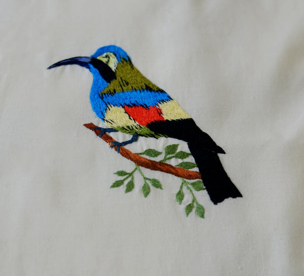Un des oiseaux colorés brodés sur le jeté de lit, celui ci est multicolore (bleu, vert, jaune,orange) et posé sur une branche avec quelques feuilles.
