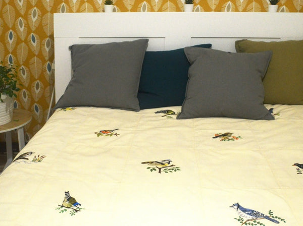 Jeté de lit couleur crème, différents types d'oiseaux colorés brodés à la main dessus, exposé sur un lit bien fait dans une chambre dont une plante sur le chevet. 