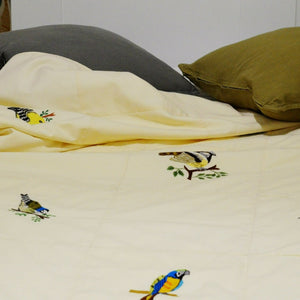 Jeté de lit couleur crème, différents types d'oiseaux colorés brodés à la main dessus, exposé sur un lit défait avec 2 coussins,